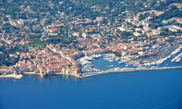 Flyfoto av St. Tropez, skrått ovenfra, på en solskinnsdag. Man ser en havn, en del sjø, og mange små bygninger samt vegetasjon.