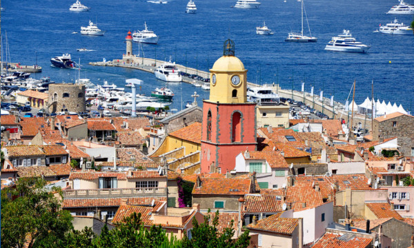 Fotografi av det berømte klokketårnet i havnen til ST. Tropez på en solskinnsdag. Underdelen er oransje-aktig, og selve toppen med klokke på er malt i en gulfarge. Rundt klokketårnet er mange små hus med oransje takstein. I det fjerne ser vi havnen med båter.